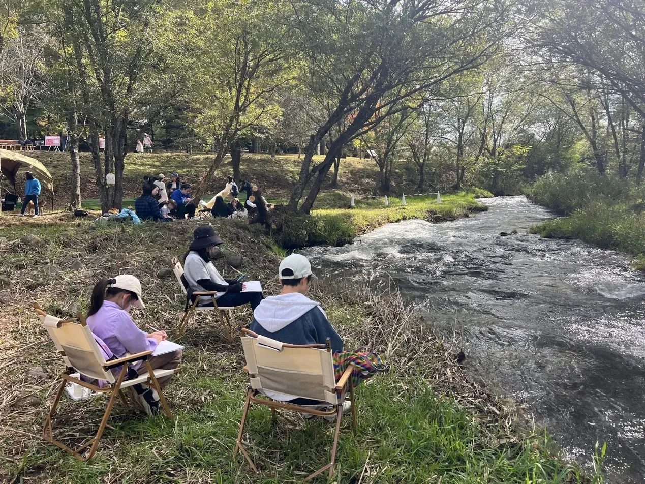 People drawing beside the Yukawa river, with more participants gathered under the trees.